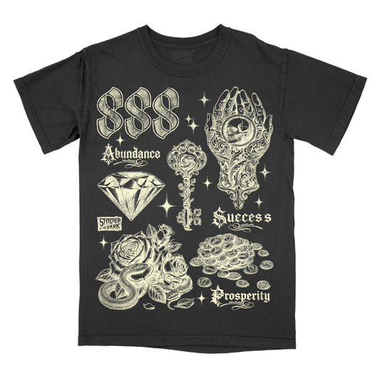 black T-shirt featuring a distressed 888 angel number design with a diamond, ornate key, mystical hand holding a skull, roses, snake, and coins, with text reading “Abundance,” “Success,” and “Prosperity,” plus Stitched in the Dark branding.