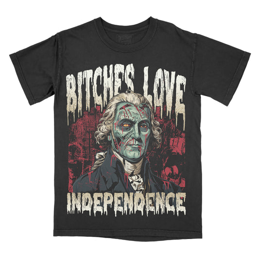 A black short-sleeve graphic t-shirt featuring a distressed print of George Washington with zombie-like green skin, the text 'BITCHES LOVE INDEPENDENCE' in a dripping white font, and a red cityscape background.