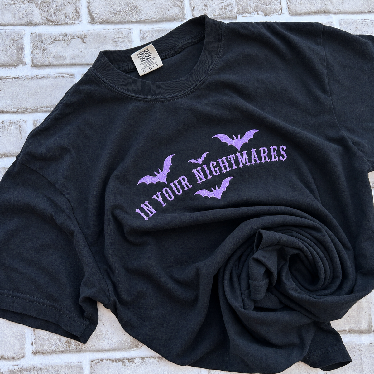 black Comfort Colors T-shirt featuring lavender embroidered gothic text reading “In Your Nightmares,” surrounded by four embroidered bats, styled on a white  background.
