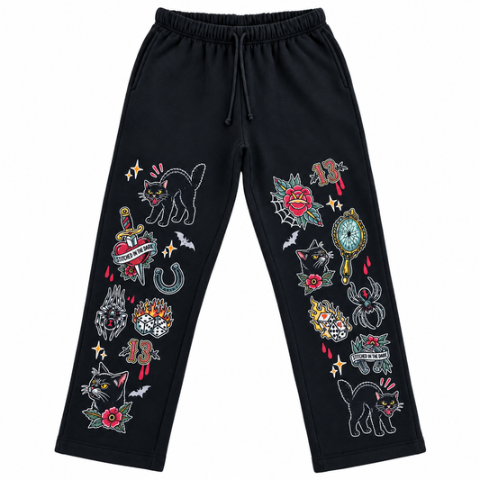 Black straight-leg sweatpants featuring colorful vintage tattoo-style graphics down both legs, including black cats, roses, bats, flaming dice, daggers, spiders, mirrors, horseshoes, number 13 motifs, and Stitched in the Dark banners.