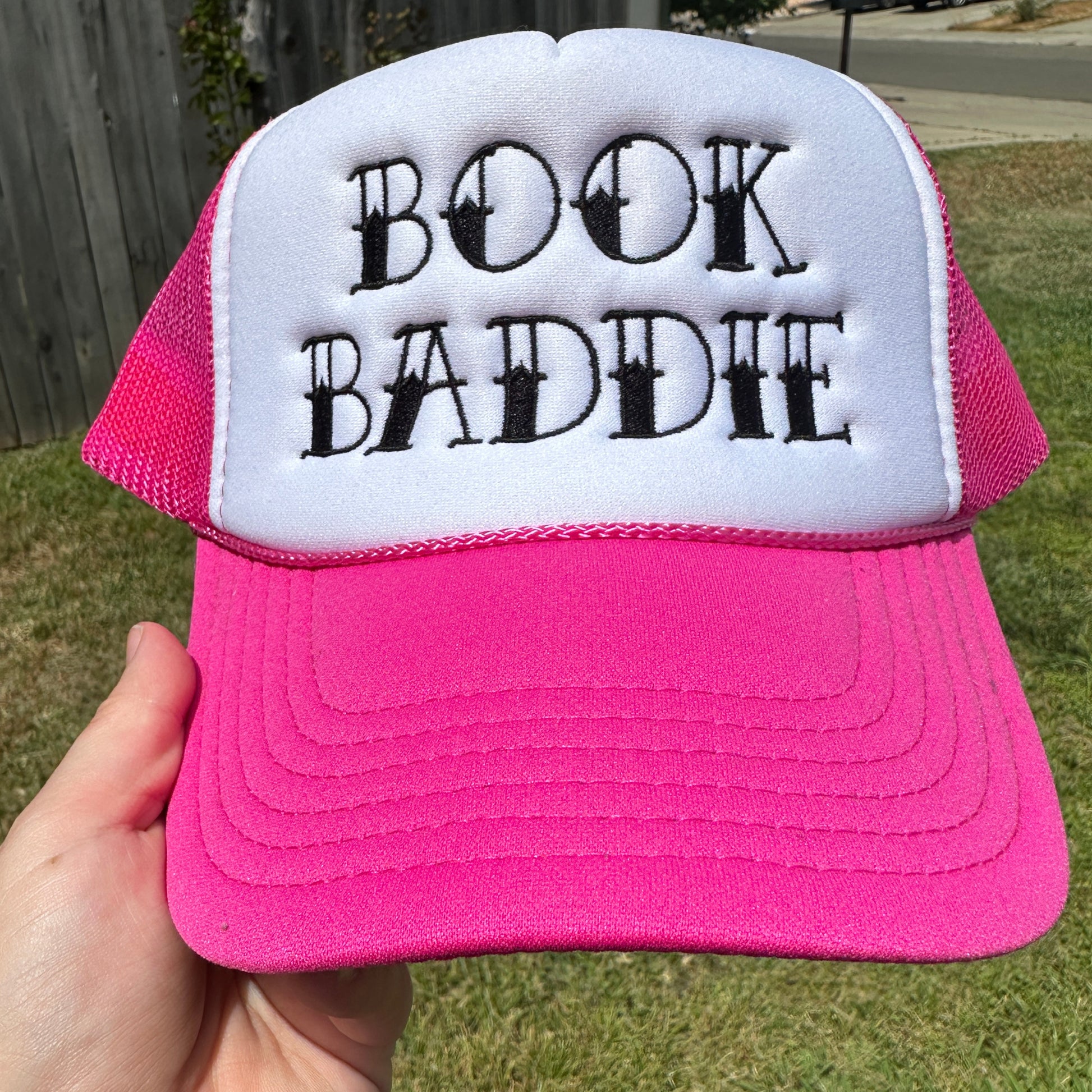 Pink and white foam trucker hat with black embroidered “Book Baddie” design, pink mesh back, pink rope detail, and curved pink bill.