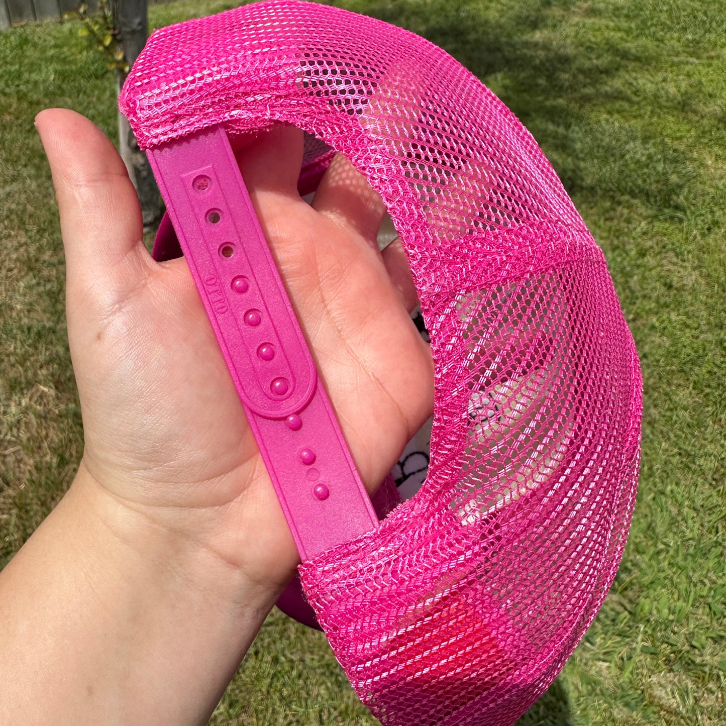 A close-up of a hand holding a bright pink trucker-style cap outdoors on a grassy lawn. The cap features a pink mesh back and a solid pink adjustable strap.