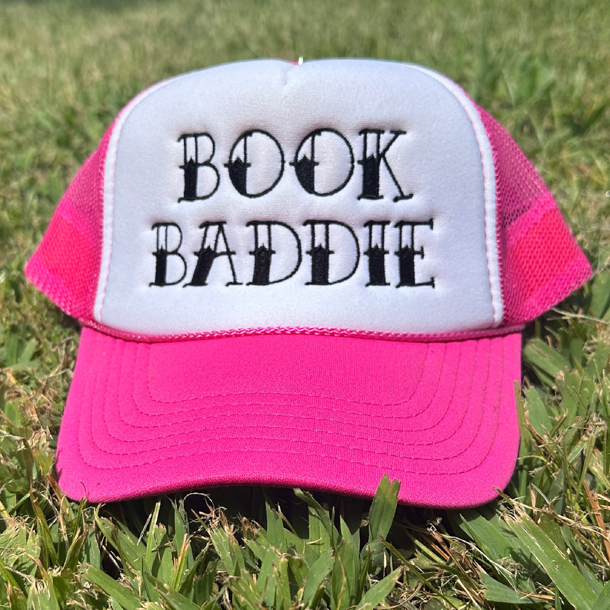 Pink and white foam trucker hat with black embroidered “Book Baddie” design, pink mesh back, pink rope detail, and curved pink bill.