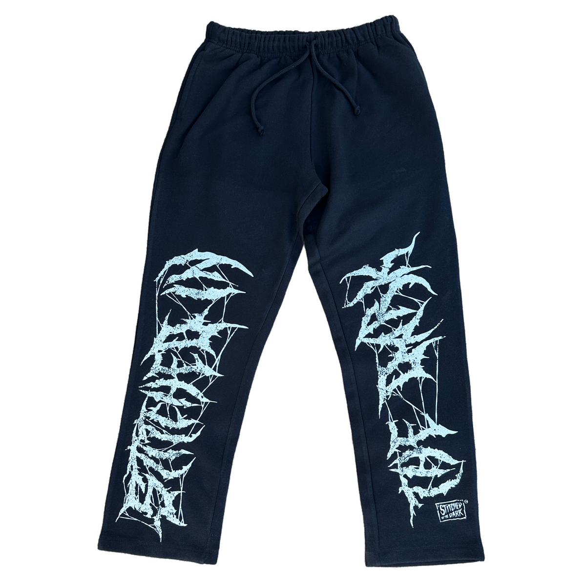 Black straight-leg sweatpants with an elastic drawstring waist featuring distressed white “Stitched in the Dark” lettering in a metal-style font printed vertically down both legs.
