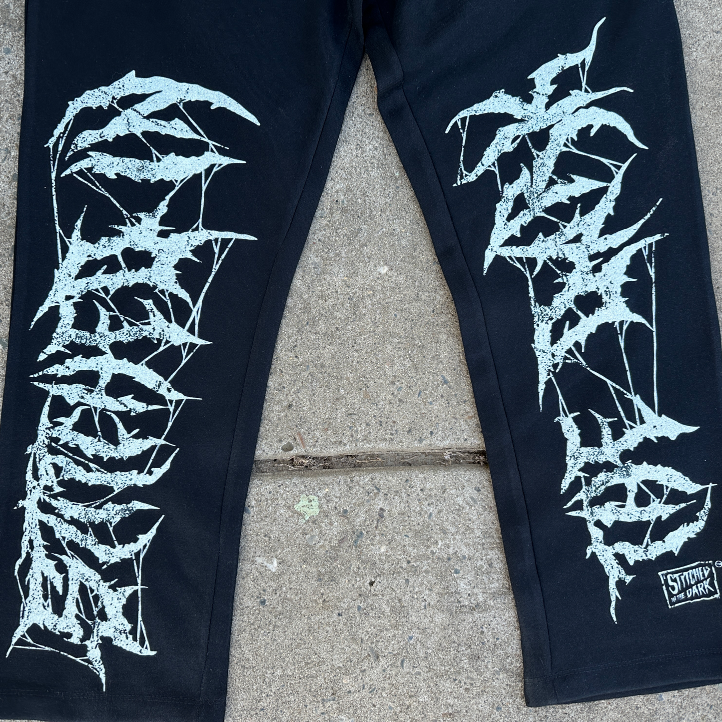 Black straight-leg sweatpants with an elastic drawstring waist featuring distressed white “Stitched in the Dark” lettering in a metal-style font printed vertically down both legs.