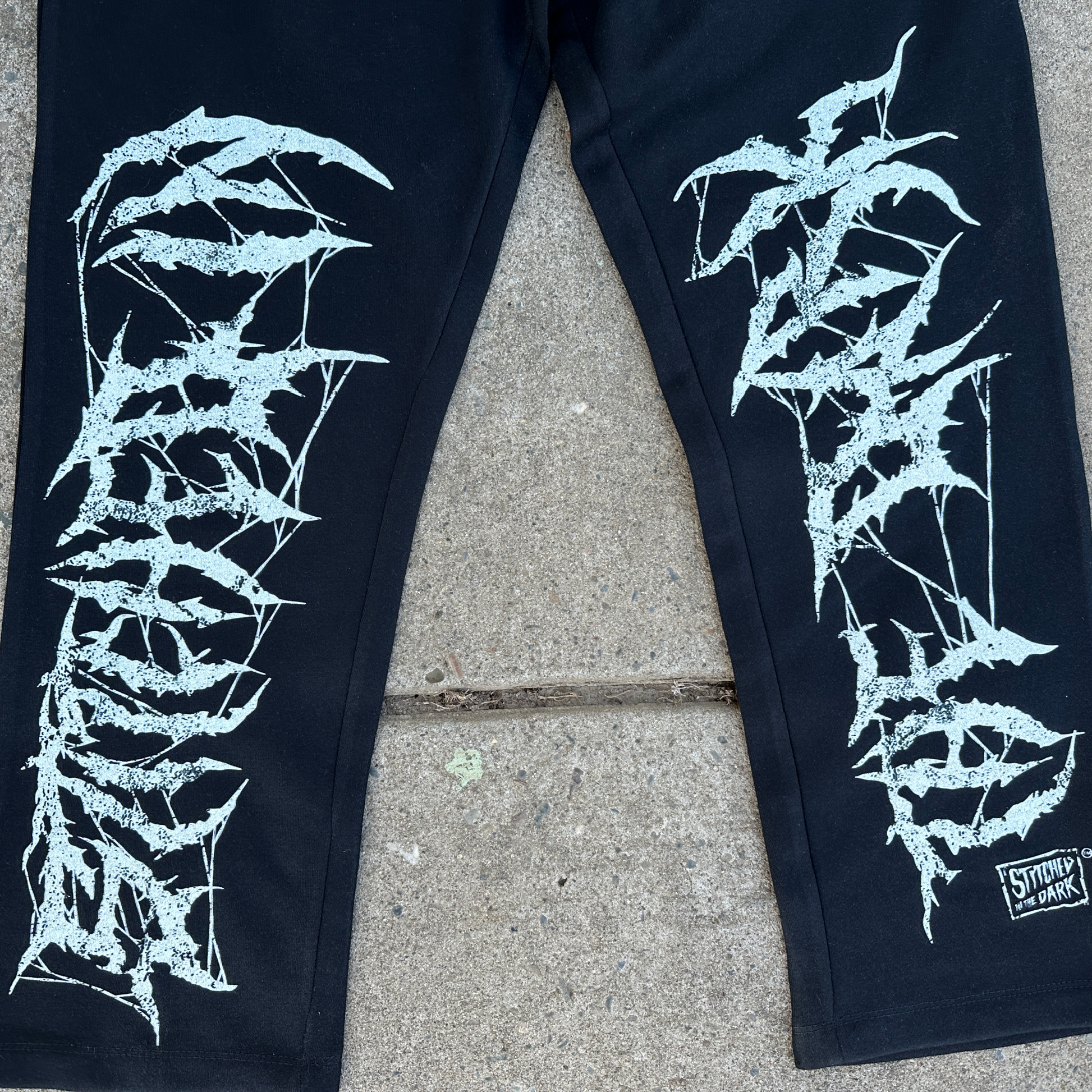 Black straight-leg sweatpants with an elastic drawstring waist featuring distressed white “Stitched in the Dark” lettering in a metal-style font printed vertically down both legs.