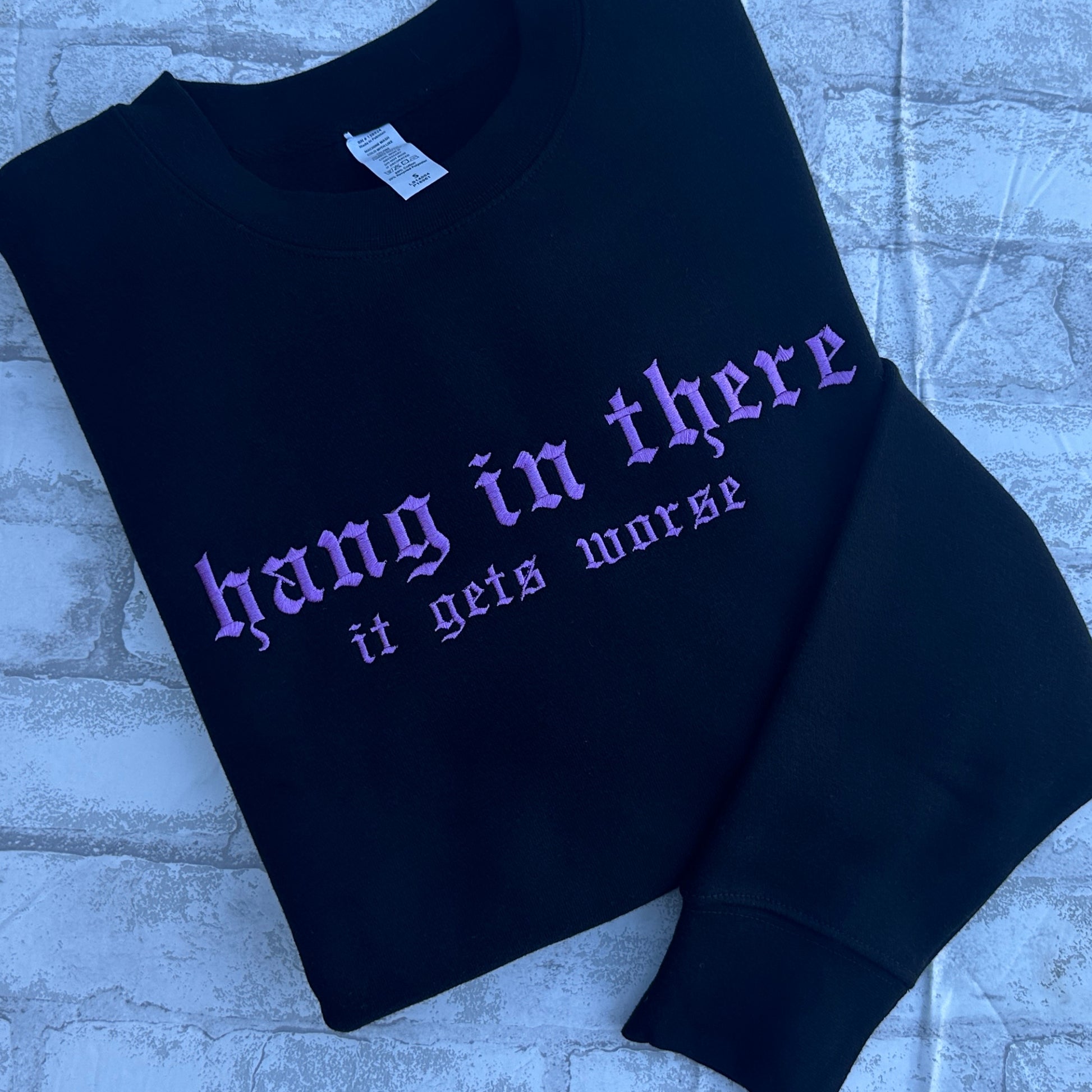 Black crewneck sweatshirt featuring lavender embroidered Gothic text that reads “hang in there” with “it gets worse” underneath, shown on a white background.