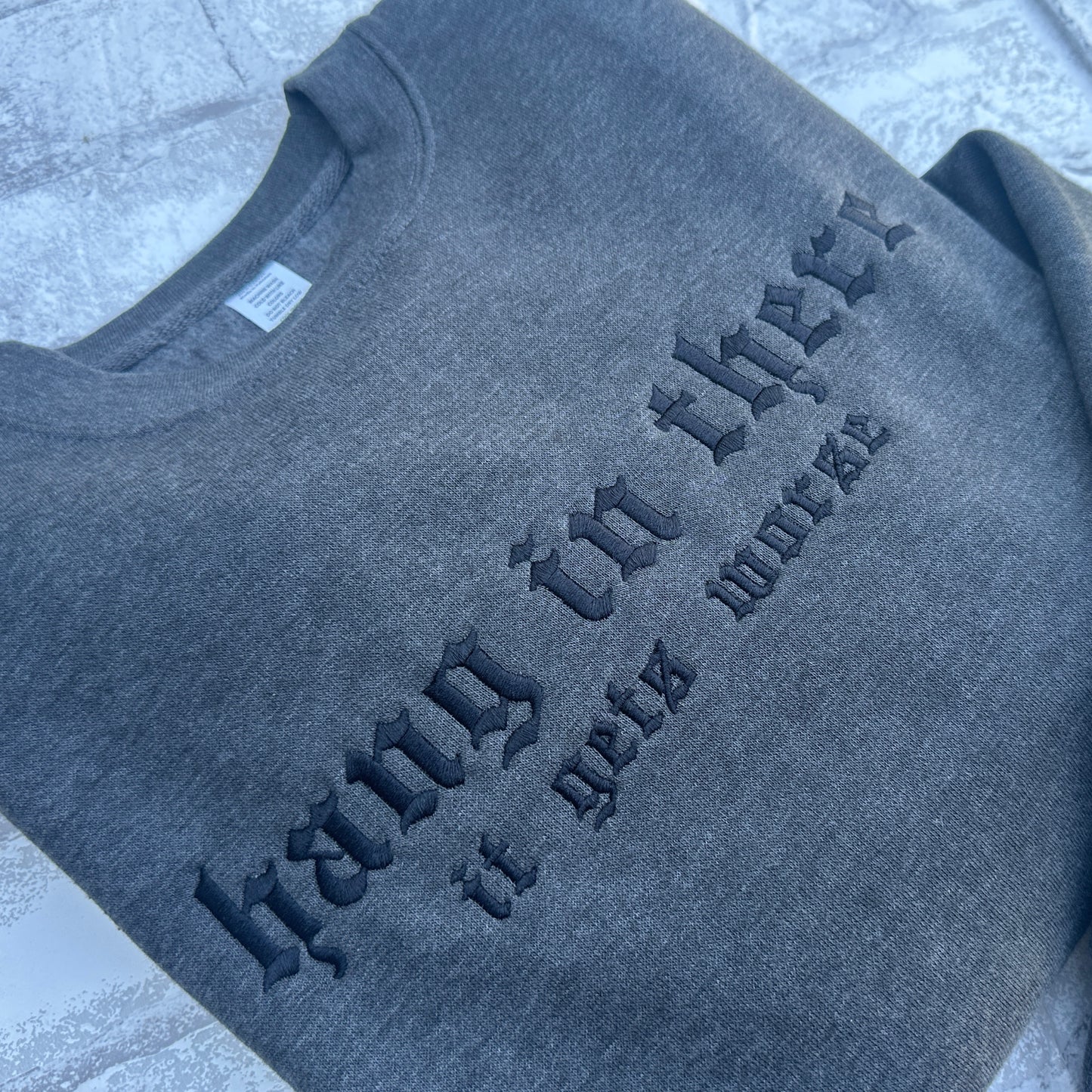 A dark gray heather crewneck sweatshirt featuring the gothic-style text 'hang in there it gets worse' in matching dark gray embroidery, laid flat on a light gray textured surface.