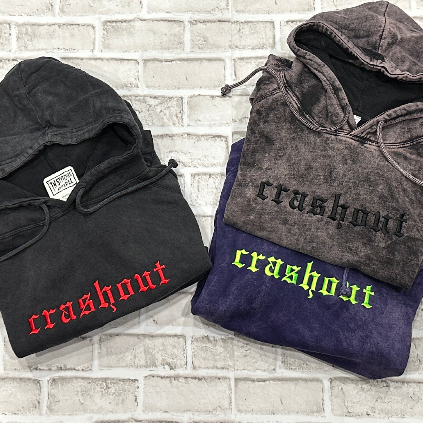 Three folded hoodies with a washed finish are arranged on a white brick background. The left hoodie is black with red 'crashout' text, and the right side shows two stacked hoodies in charcoal and purple with black and neon green 'crashout' text respectively.