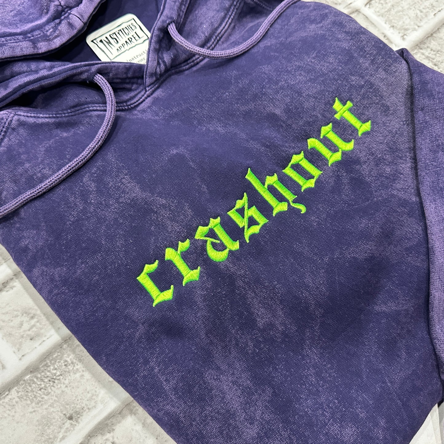 A close-up of a purple mineral-wash hoodie featuring the word 'crashout' embroidered in a bright neon green gothic font across the chest. 