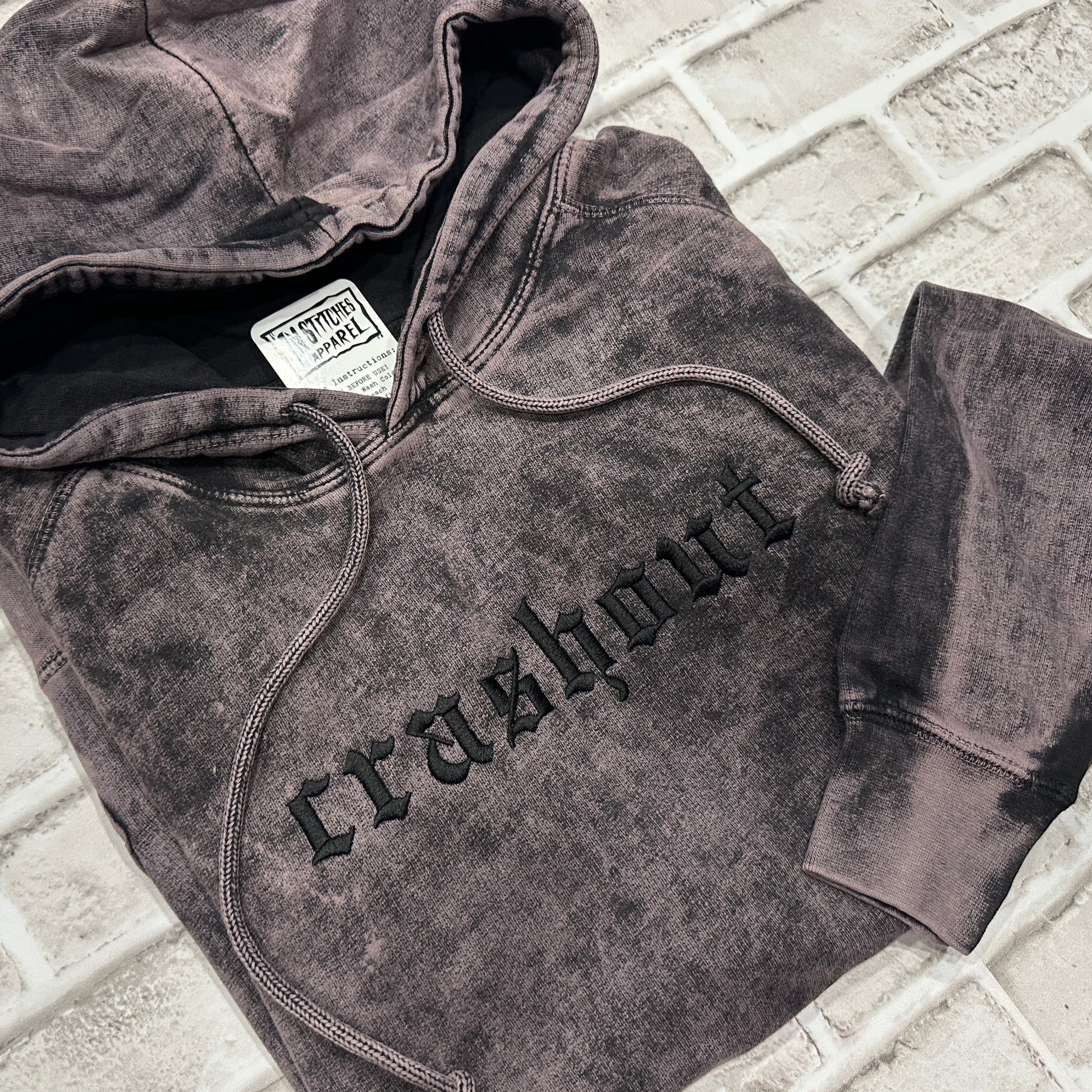 A dark gray acid-wash pullover hoodie with 'crashout' embroidered in black gothic lettering across the chest, laid flat on a light gray brick surface.