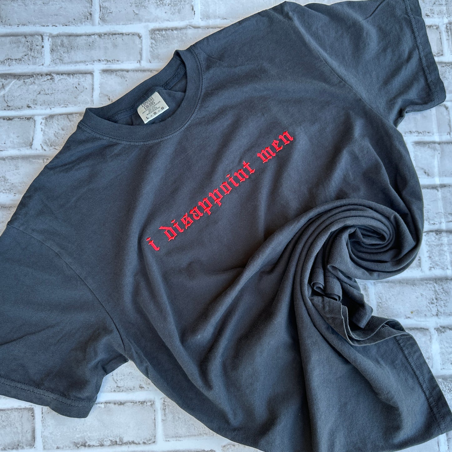 A black short-sleeve t-shirt with the phrase 'i disappoint men' embroidered across the chest in a red gothic font, laid flat against a white background.
