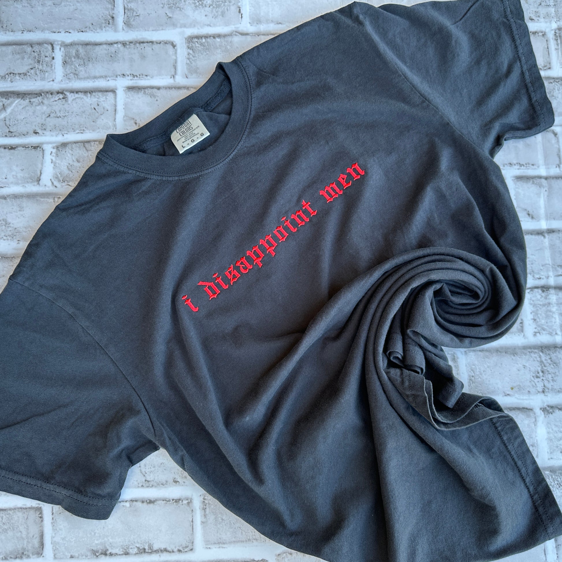 A black short-sleeve t-shirt with the phrase 'i disappoint men' embroidered across the chest in a red gothic font, laid flat against a white background.