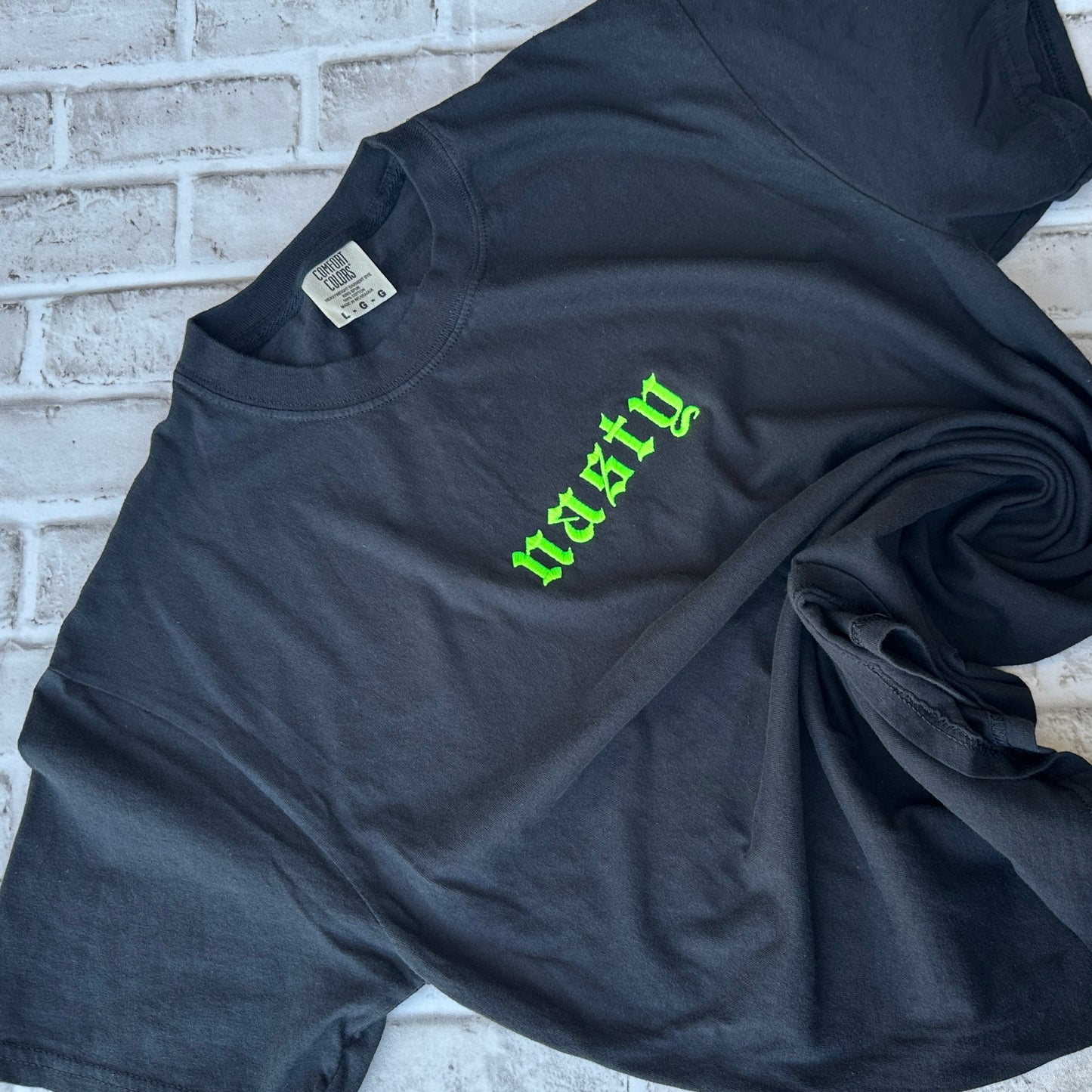 A black short-sleeve t-shirt with the word 'nasty' embroidered in a bright green gothic font across the chest, displayed flat against a white background.