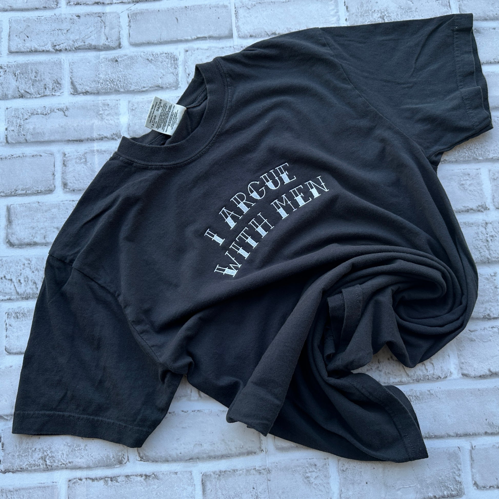A black short-sleeve t-shirt with the phrase 'I ARGUE WITH MEN' embroidered in white arched text across the chest, laid flat on a white background.