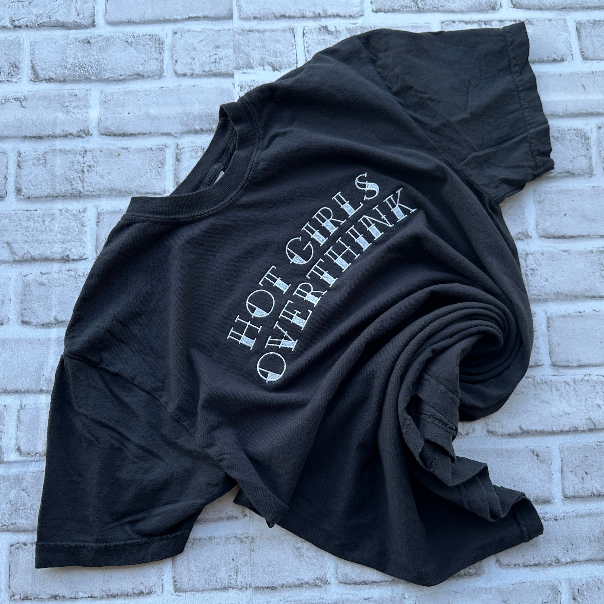 A black short-sleeve t-shirt with the phrase 'HOT GIRLS OVERTHINK' embroidered in a white, stylized font across the chest, displayed flat against a white background.