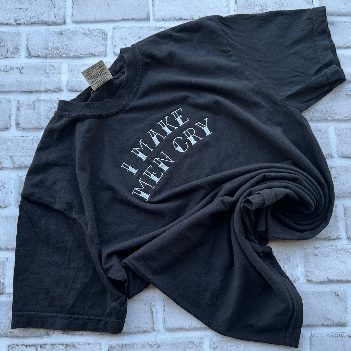 A black short-sleeve t-shirt with the phrase 'I MAKE MEN CRY' embroidered in a distressed white font across the chest, laid flat against a white background.