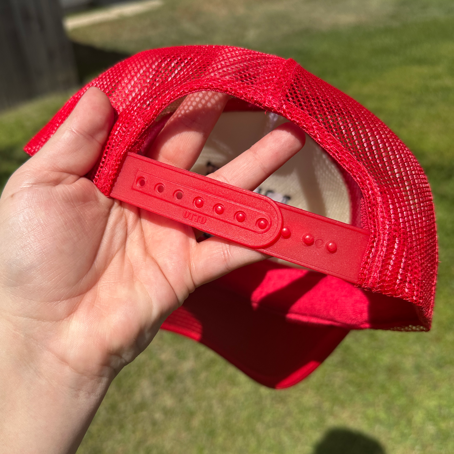 A close-up of a person's hand holding the back strap of a red mesh trucker hat outdoors on a grassy lawn.