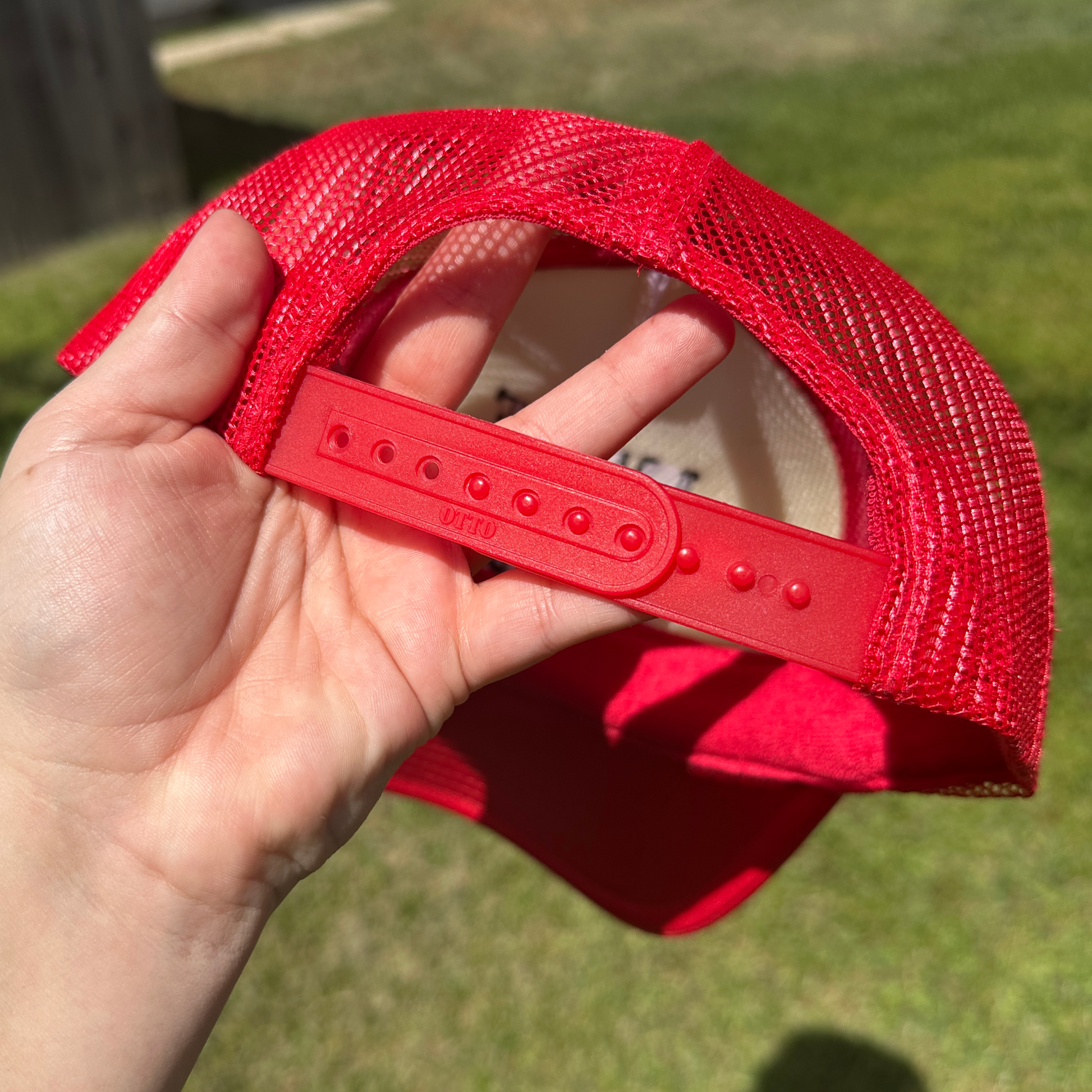 A close-up of a person's hand holding the back strap of a red mesh trucker hat outdoors on a grassy lawn.