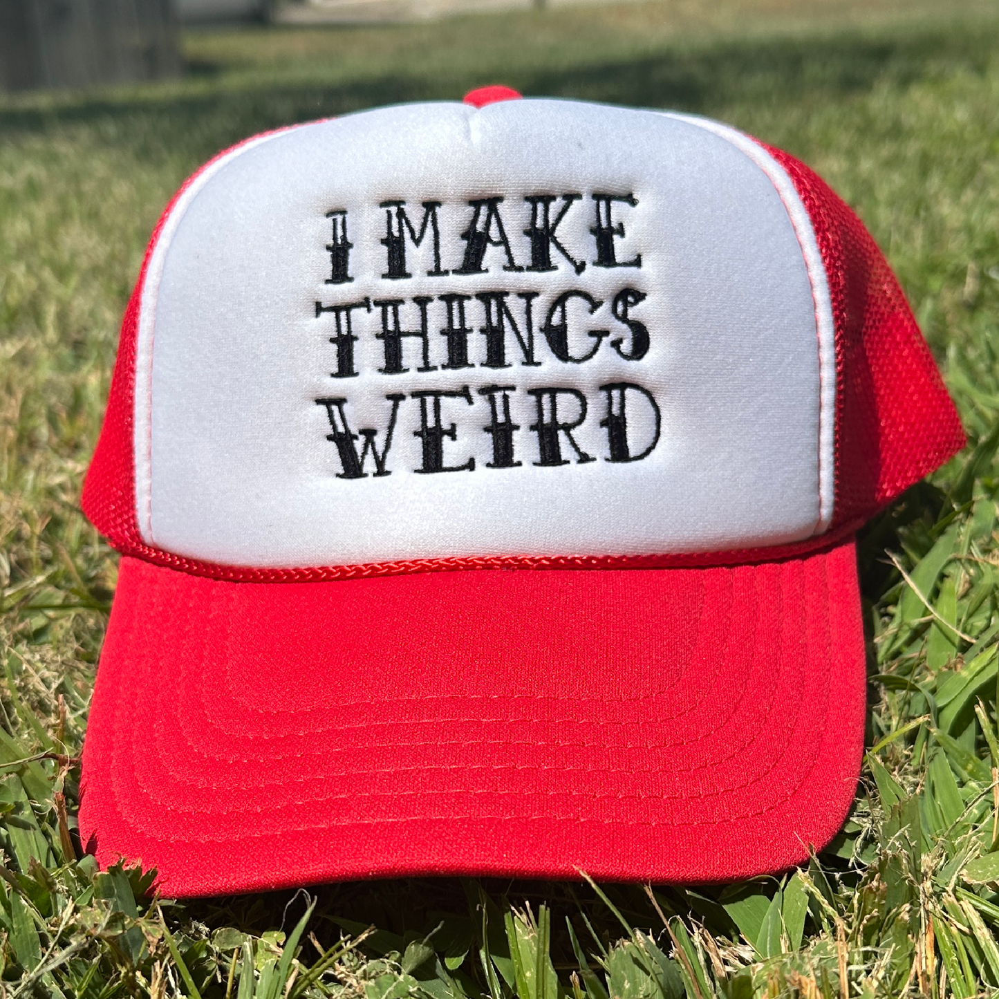 A red and white trucker hat with the embroidered text 'I MAKE THINGS WEIRD' on the front, resting on green grass outdoors.