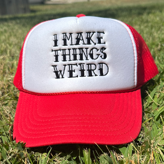 A red and white trucker hat with the embroidered text 'I MAKE THINGS WEIRD' on the front, resting on green grass outdoors.
