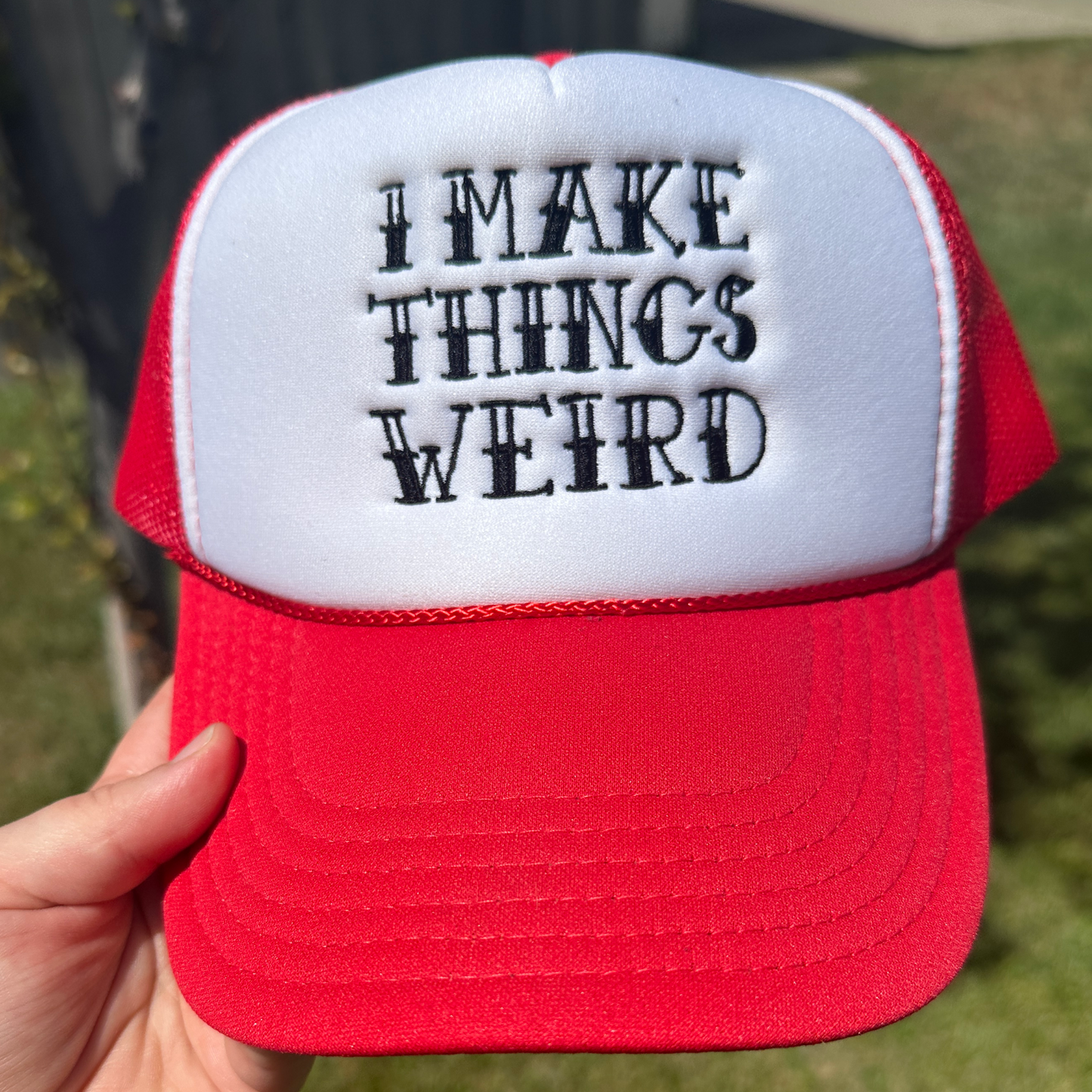 A red and white trucker hat held by a hand, featuring the text 'I MAKE THINGS WEIRD' embroidered in black on the white front panel. The background shows a blurred outdoor setting with green grass.
