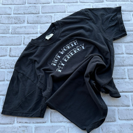 A black graphic t-shirt with the text 'NOT WORTH MY ENERGY' embroidered in white arched lettering, laid flat on a white painted brick wall.