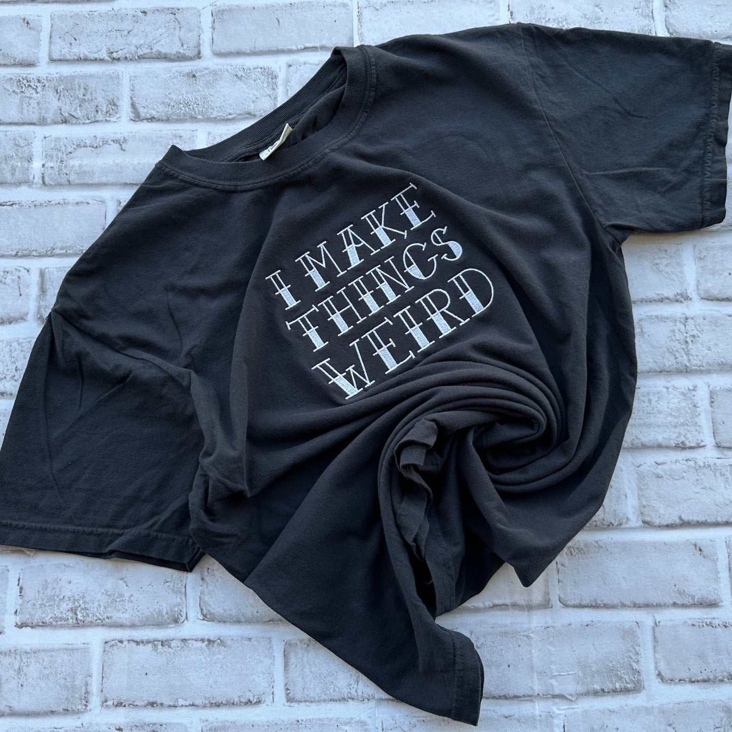 A black short-sleeve t-shirt with the phrase 'I MAKE THINGS WEIRD' embroidered in a stylized white font on the front, laid flat against a white painted brick wall.