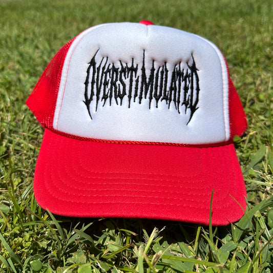 A red and white trucker hat with the word 'OVERSTIMULATED' in a black gothic font embroidered on the front, resting on green grass.
