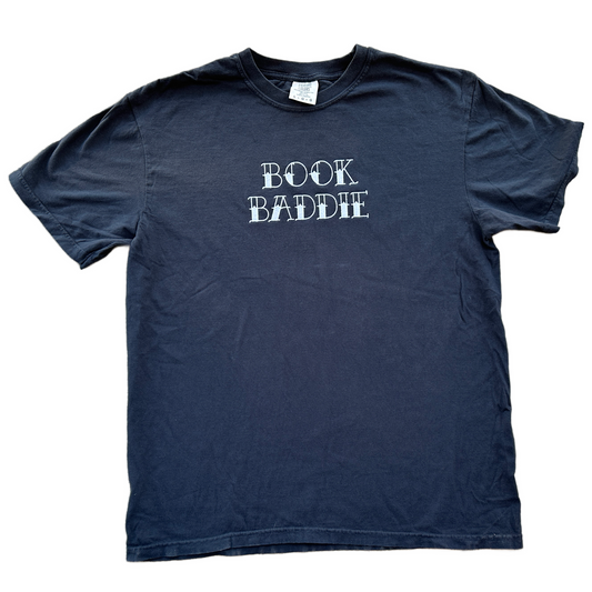 A black short-sleeve t-shirt with the phrase 'BOOK BADDIE' embroidered in a distressed white font across the chest, laid flat against a white background.