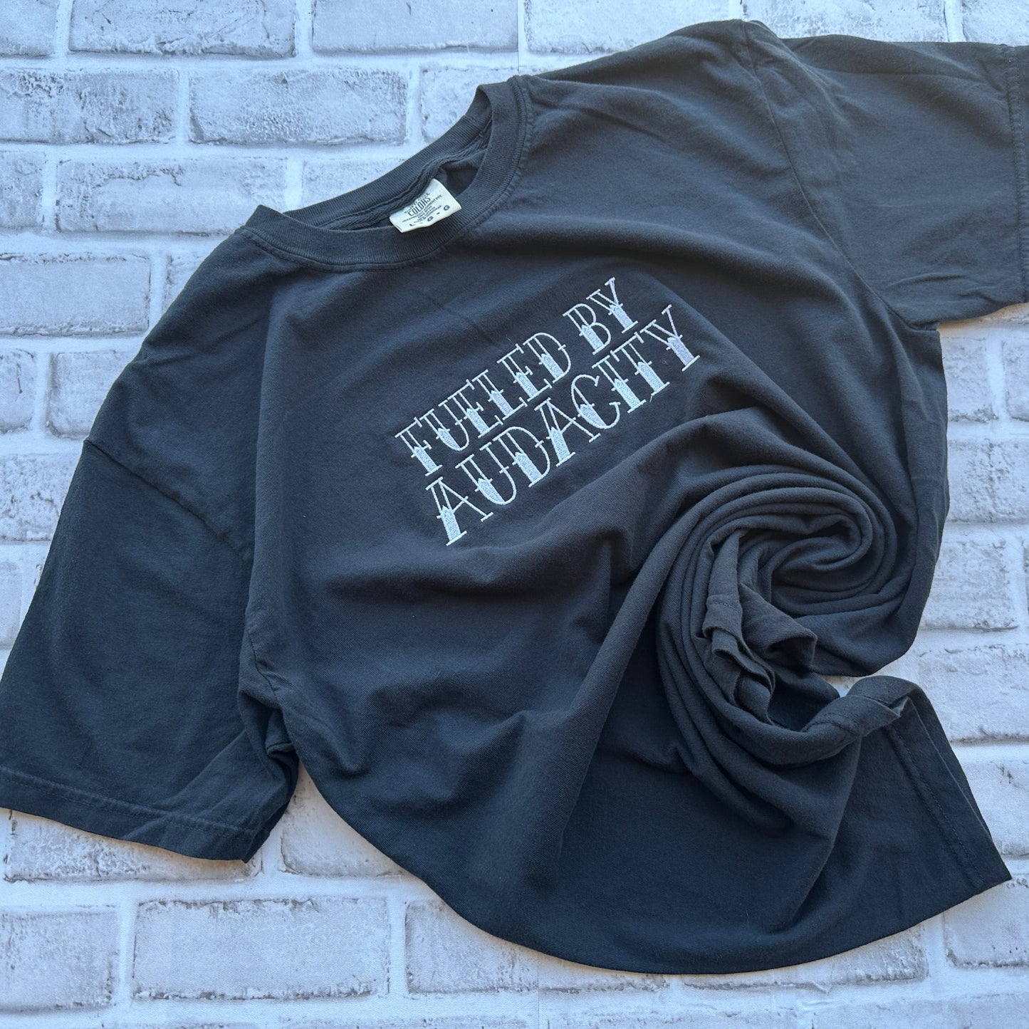 A black short-sleeve t-shirt with the phrase 'FUELED BY AUDACITY' embroidered in a distressed white font across the chest, displayed flat against a white background.