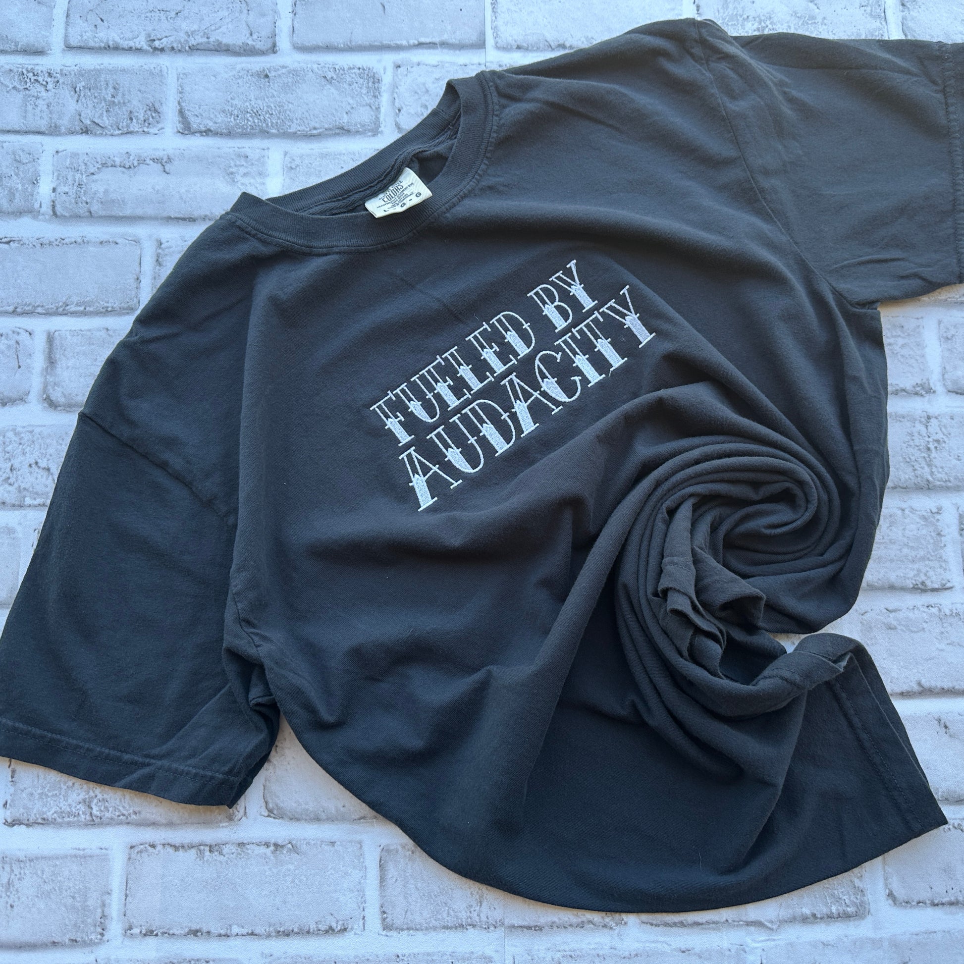 A black short-sleeve t-shirt with the phrase 'FUELED BY AUDACITY' embroidered in a distressed white font across the chest, displayed flat against a white background.