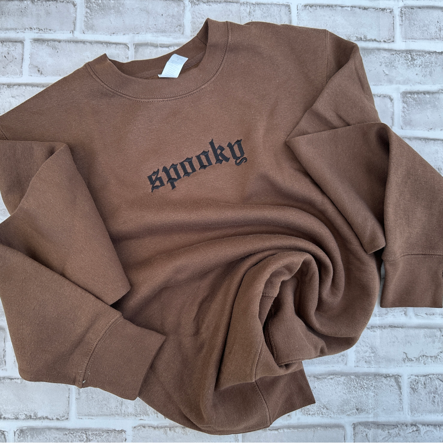 Brown crewneck sweatshirt featuring black embroidered Old English text across the chest that reads “spooky,” shown on a white background.
