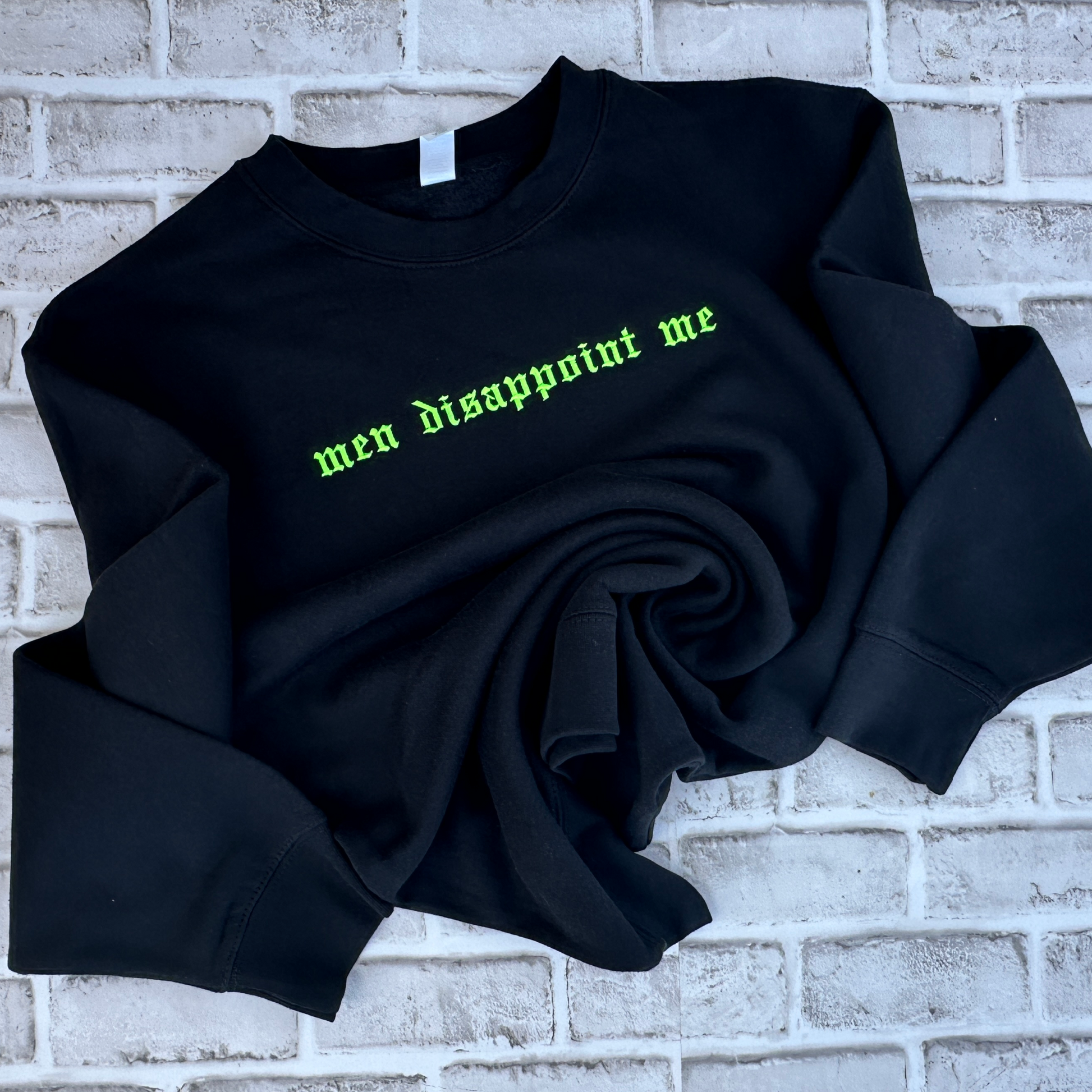 Black crewneck sweatshirt featuring neon green embroidered Old English text across the chest that reads “men disappoint me,” shown on a white background.
