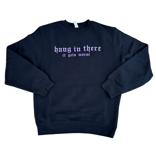 Black crewneck sweatshirt featuring lavender embroidered Gothic text that reads “hang in there” with “it gets worse” underneath, shown on a white background.