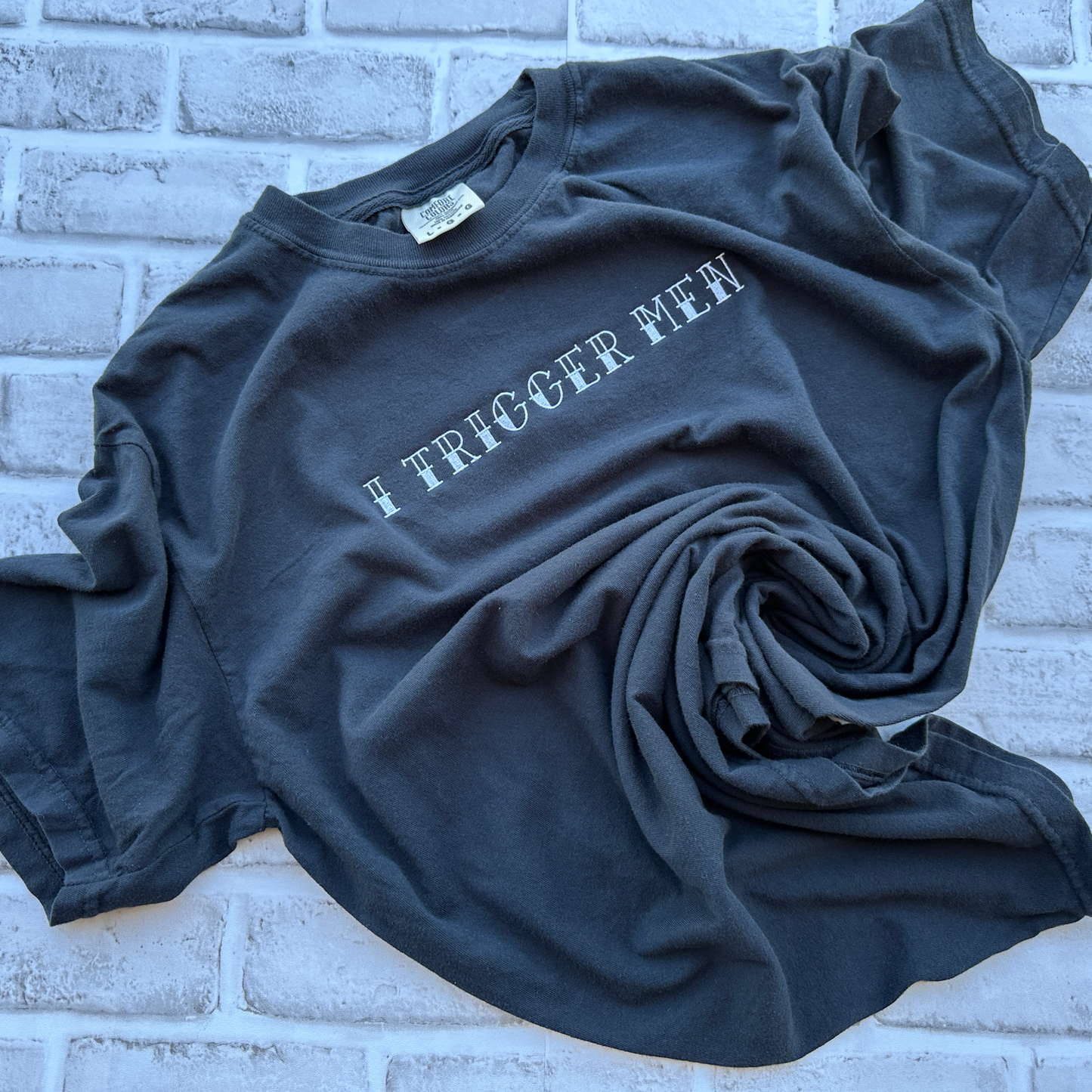 A black short-sleeve crewneck t-shirt with the text 'I TRIGGER MEN' embroidered in white across the chest, displayed flat against a white background.
