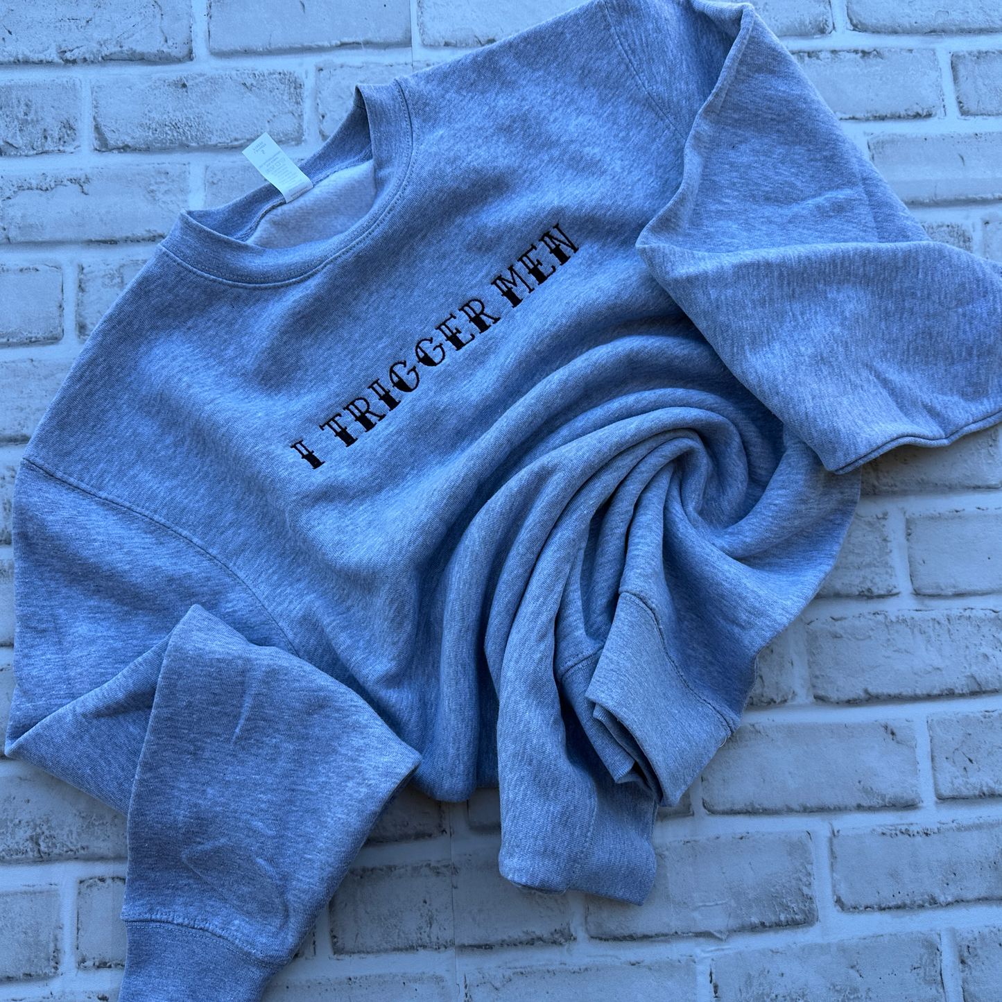 A heather grey crewneck sweatshirt with the text 'I TRIGGER MEN' embroidered in black across the chest, laid flat on a white background.