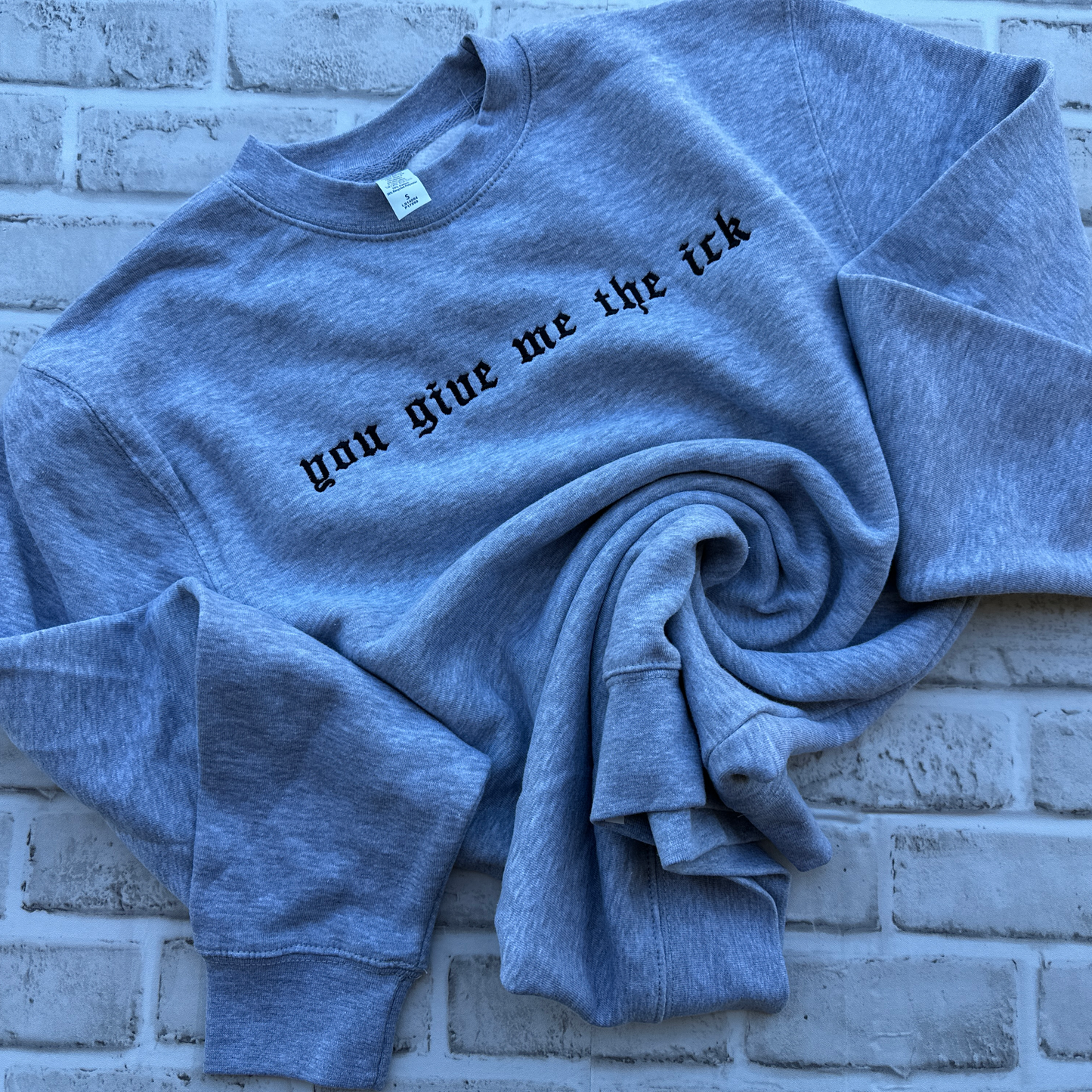 A heather grey crewneck sweatshirt with the phrase 'you give me the ick' embroidered across the chest in a black gothic font, displayed flat against a white background.