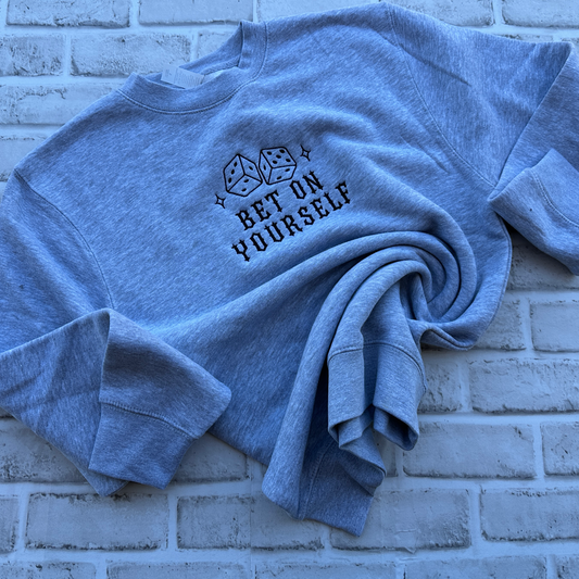 A grey crewneck sweatshirt with a black graphic of two dice and the text 'BET ON YOURSELF' embroidered on the chest, displayed flat against a white background.