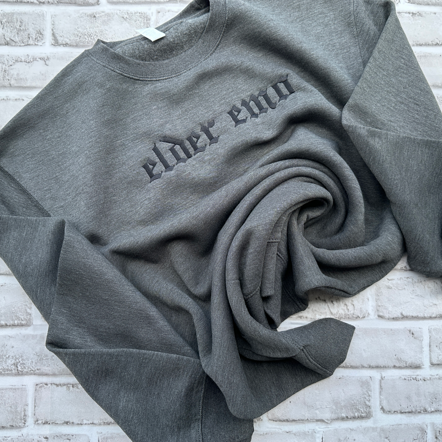 Charcoal gray crewneck sweatshirt featuring black embroidered Old English text across the chest that reads “elder emo.