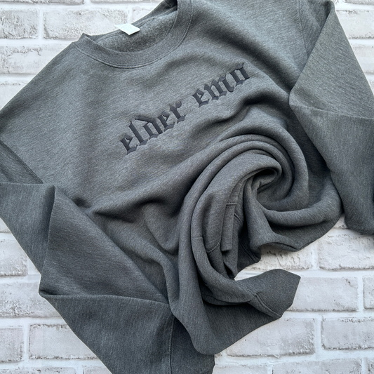 Charcoal gray crewneck sweatshirt featuring black embroidered Old English text across the chest that reads “elder emo.