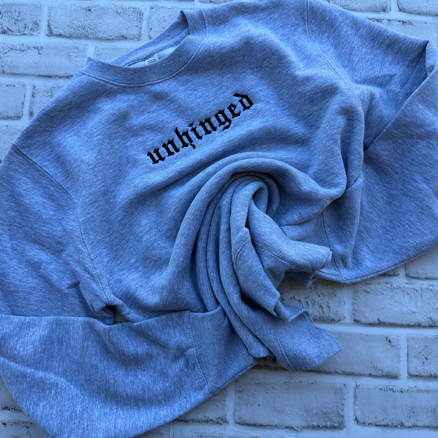 A heather gray crewneck sweatshirt with the word 'unhinged' embroidered in a black gothic font across the chest, displayed flat against a solid white background.