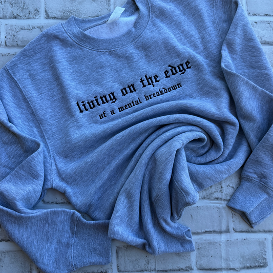 A heather gray crewneck sweatshirt with black gothic text reading 'living on the edge of a mental breakdown' across the chest, displayed flat against a white background.