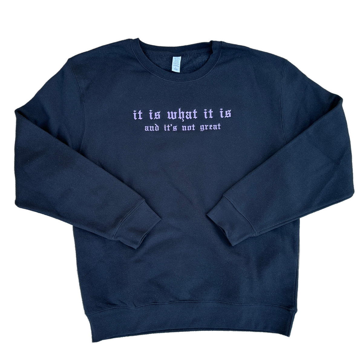 Black crewneck sweatshirt featuring purple embroidered Old English text across the chest that reads “it is what it is” with smaller text underneath reading “and it’s not great.”