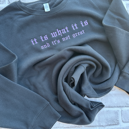 Black crewneck sweatshirt featuring purple embroidered Old English text across the chest that reads “it is what it is” with smaller text underneath reading “and it’s not great.”