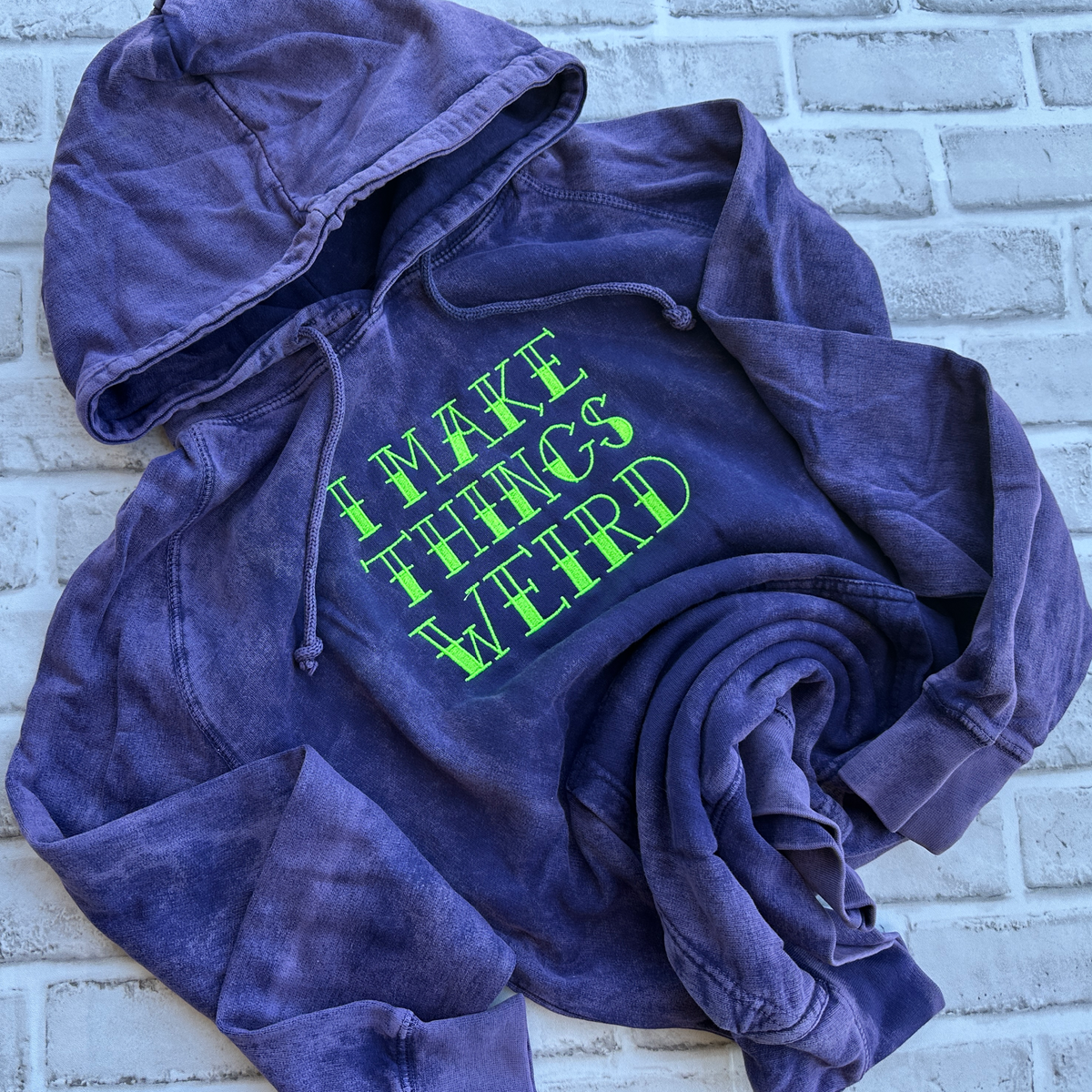 Purple mineral-washed hoodie featuring bright neon green embroidered text across the chest that reads “I MAKE THINGS WEIRD,” with a drawstring hood and front kangaroo pocket.