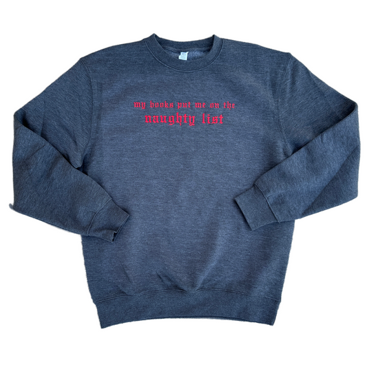 Charcoal gray crewneck sweatshirt featuring bright red embroidered Old English text across the chest that reads “my books put me on the naughty list.