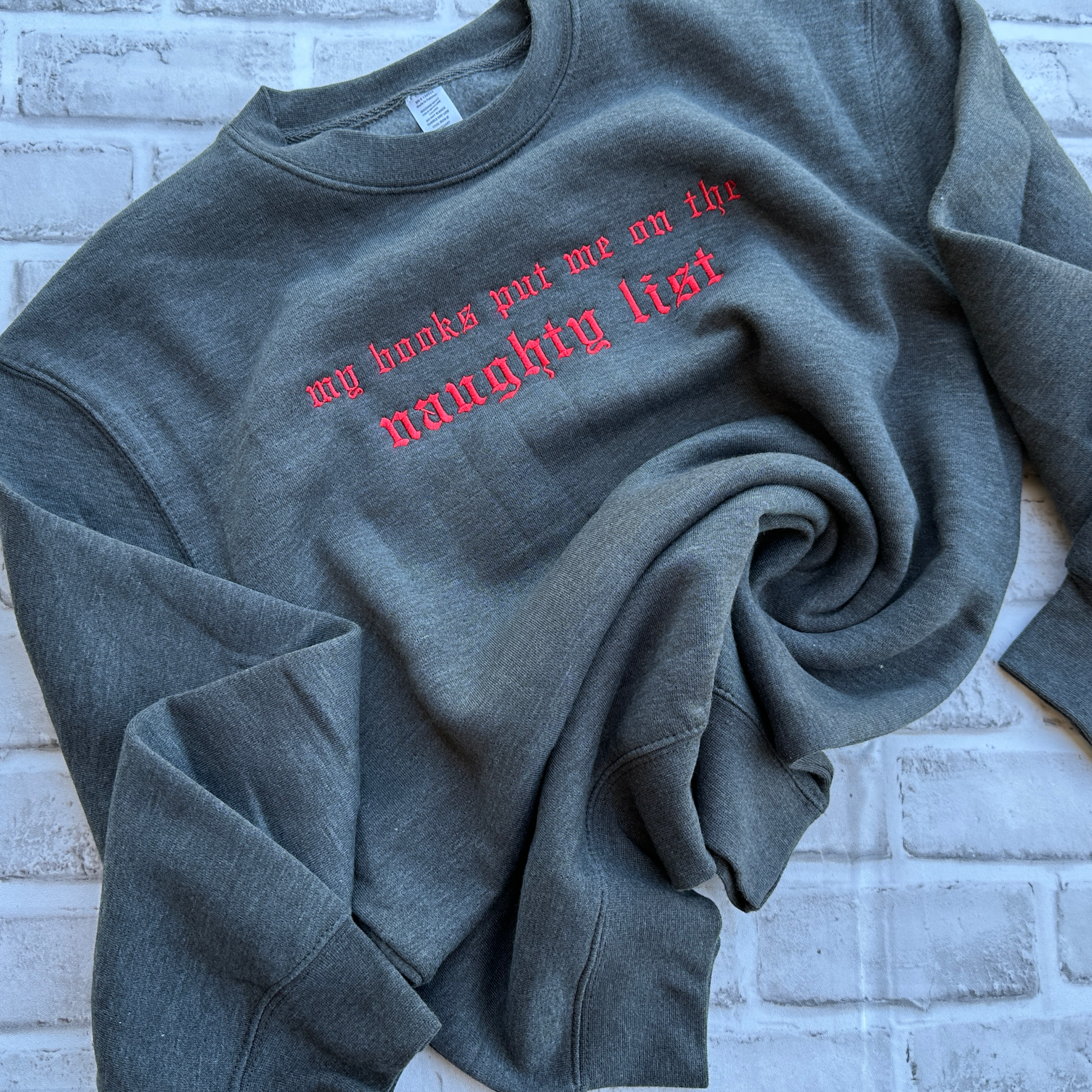 Charcoal gray crewneck sweatshirt featuring bright red embroidered Old English text across the chest that reads “my books put me on the naughty list.