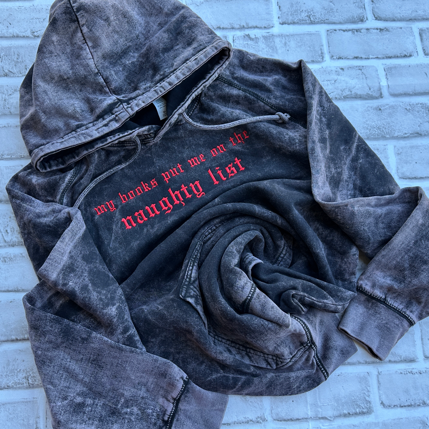 Vintage cloud black mineral-washed hoodie featuring bright red embroidered Old English text across the chest that reads “my books put me on the naughty list,” with a drawstring hood and front kangaroo pocket.