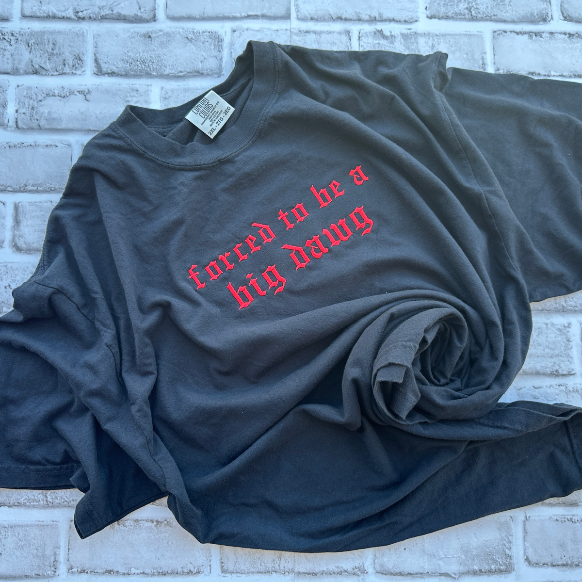 Black Comfort Colors T-shirt featuring red Old English embroidered text across the chest that reads “forced to be a big dawg,” shown flat on a white background.